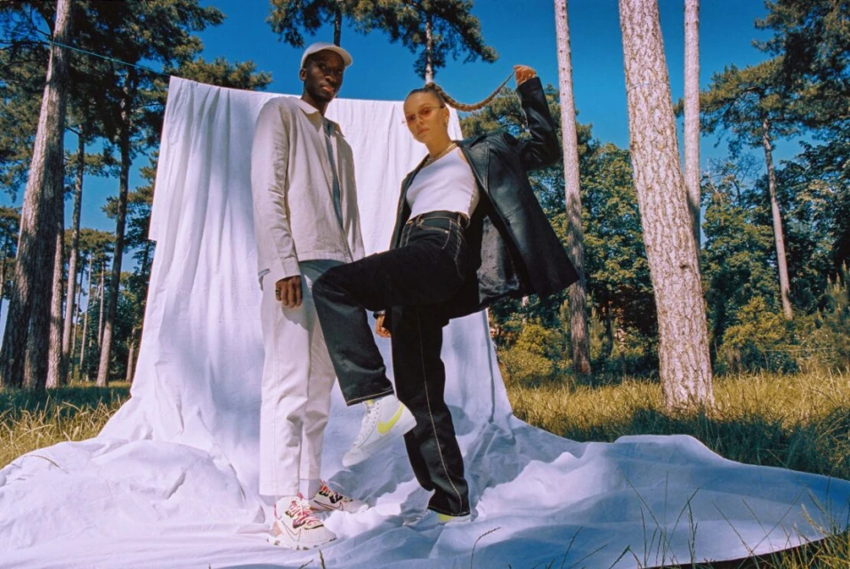 Two models pose in an outdoor fashion shot, wearing a jacket, white shirt, trousers, and sneakers, against a backdrop of trees.