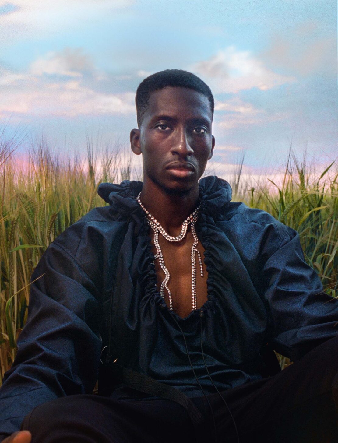 Dark-skinned man in a blue silky top with an extravagant diamond necklace poses outdoors in a wheat field under a sky with pink clouds.