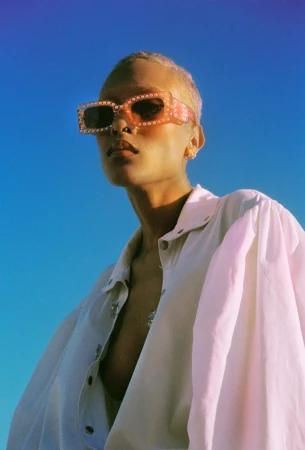 Model wearing embellished sunglasses and a white collared shirt against a bright blue sky background.