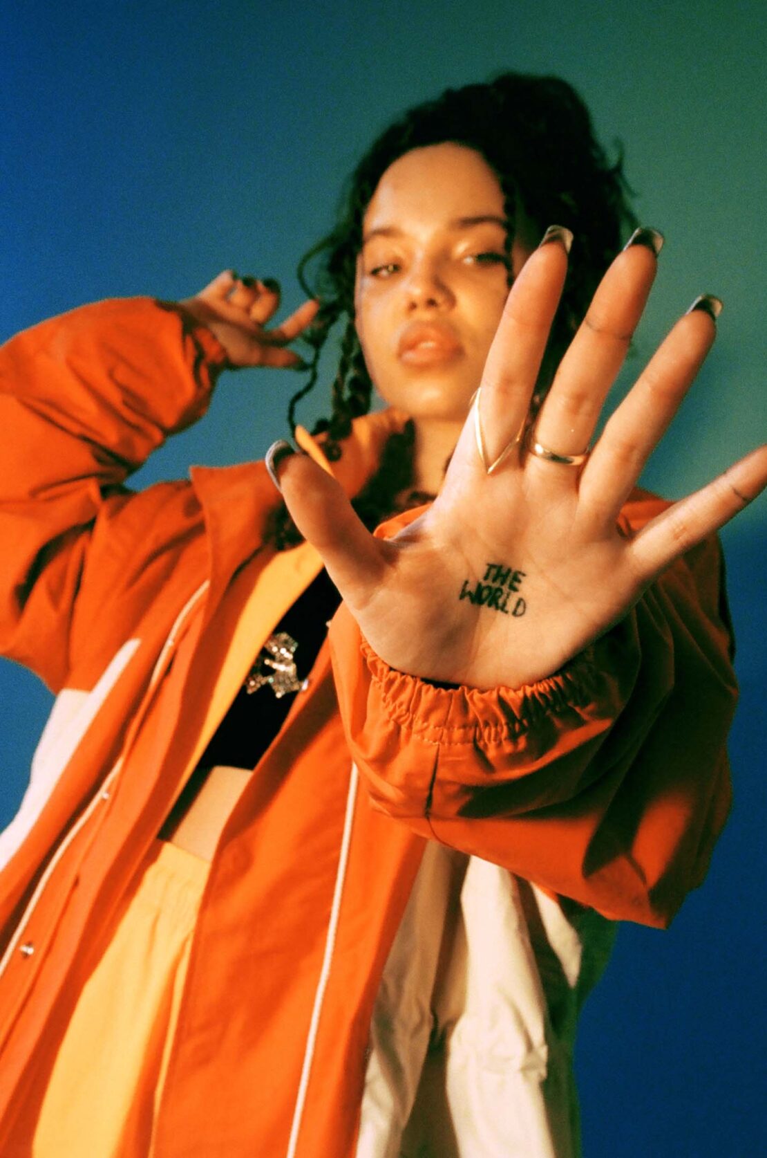 Young woman in an orange jacket presents her hand, adorned with a ring and 'The World' tattoo, against a gradient blue backdrop.