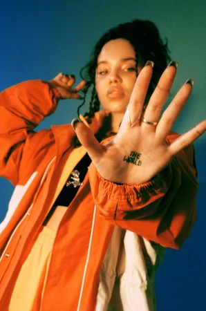 Young woman in an orange jacket presents her hand, adorned with a ring and 'The World' tattoo, against a gradient blue backdrop.