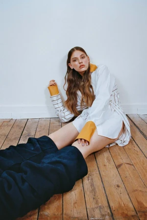 Woman with long brown hair seated on a wooden floor wearing a white shirt with yellow cuffs and dark trousers