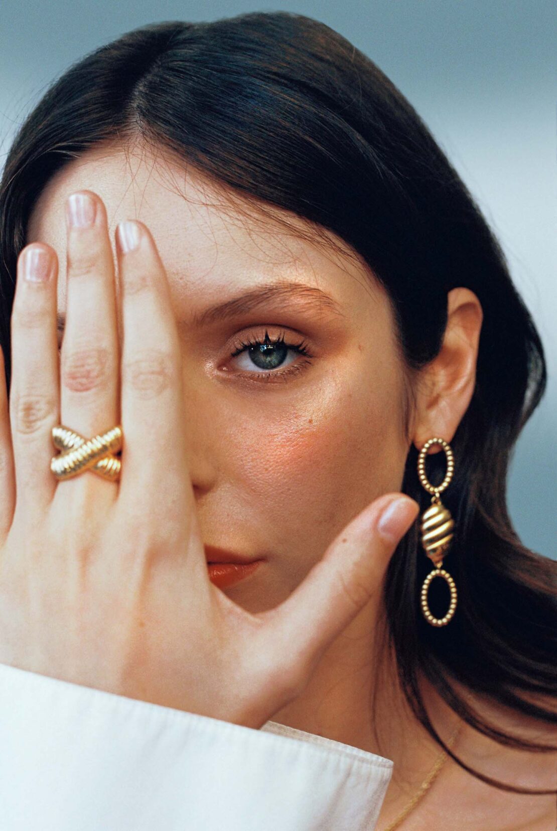 Close-up of a woman with gold hoop earrings and a criss-cross ring covering half her face, showcasing beauty and fashion accessories.