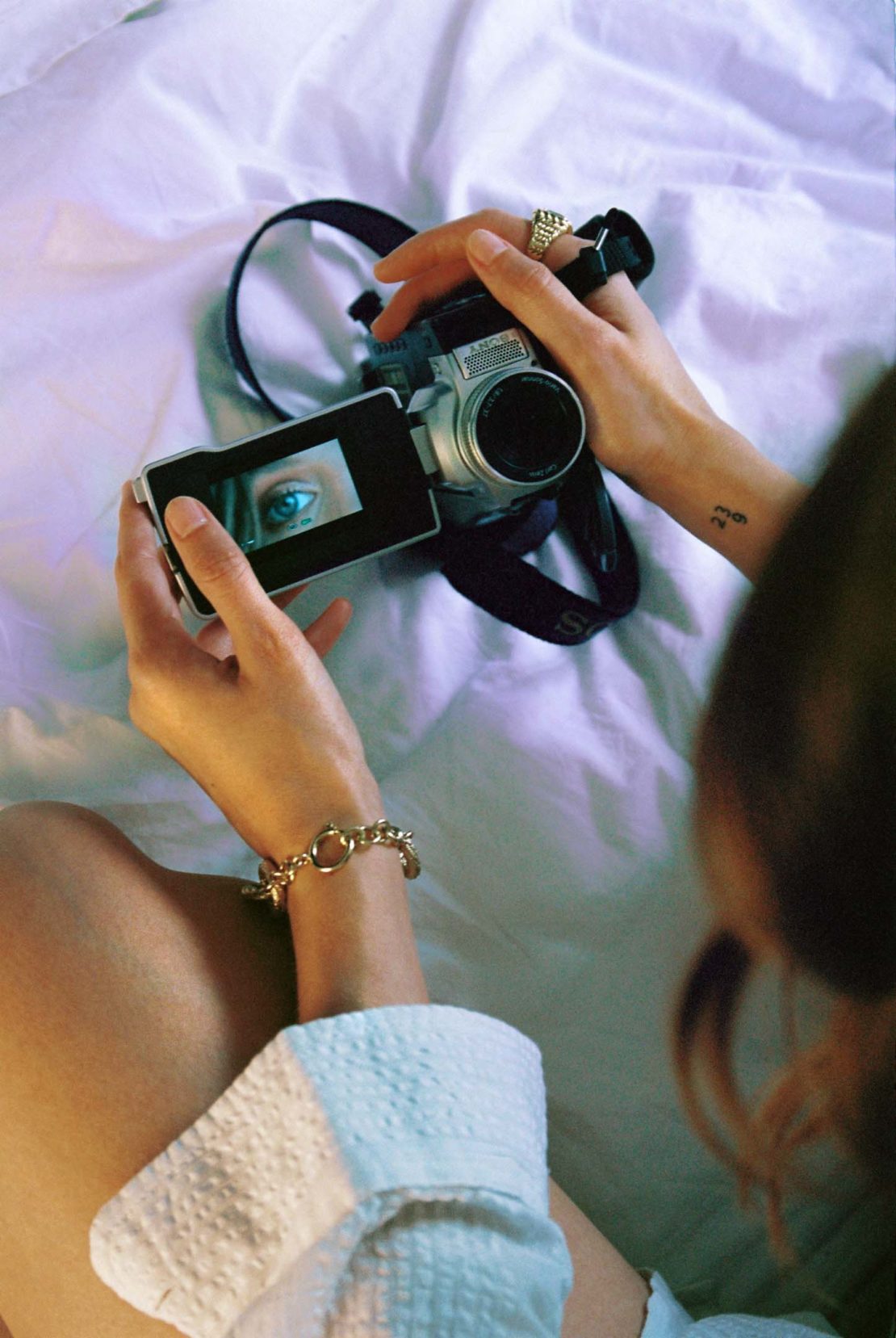 Woman holds silver video camera in a close-up, angled shot on a white bed, showing the camera screen reflecting an eye.