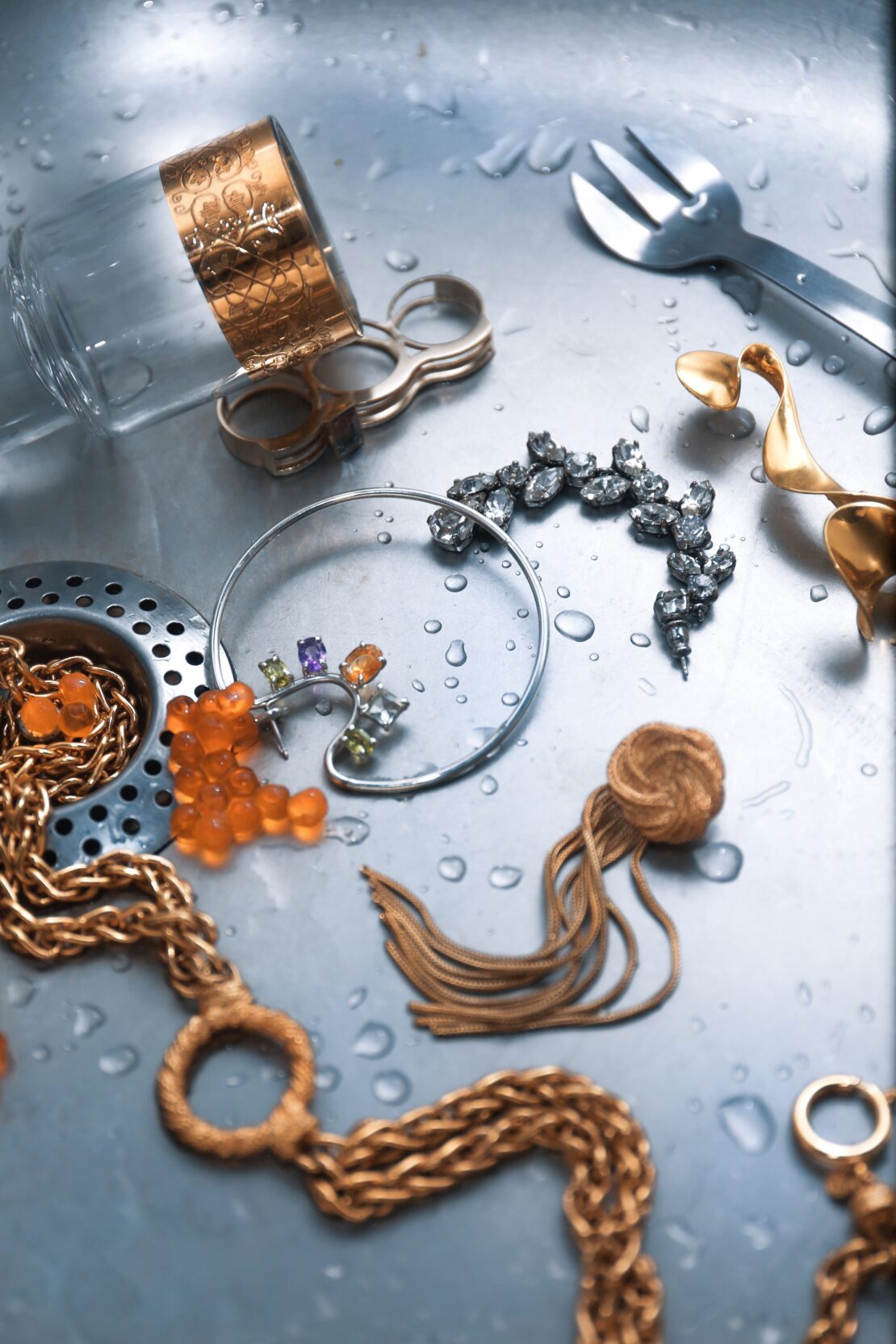 High-angle view of jewellery and tableware in a wet stainless steel kitchen sink, featuring amber and silver jewellery, glass, and silverware.