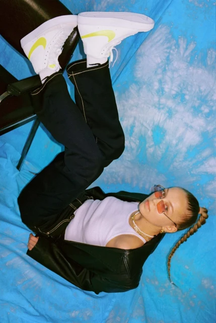 A young woman poses on a blue backdrop with Nike high-top sneakers, black pants, and a white tank top and black jacket.