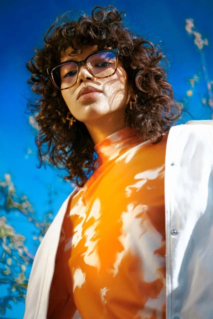 A young adult woman with curly hair poses wearing glasses and a patterned orange top against a blue floral background in a portrait.