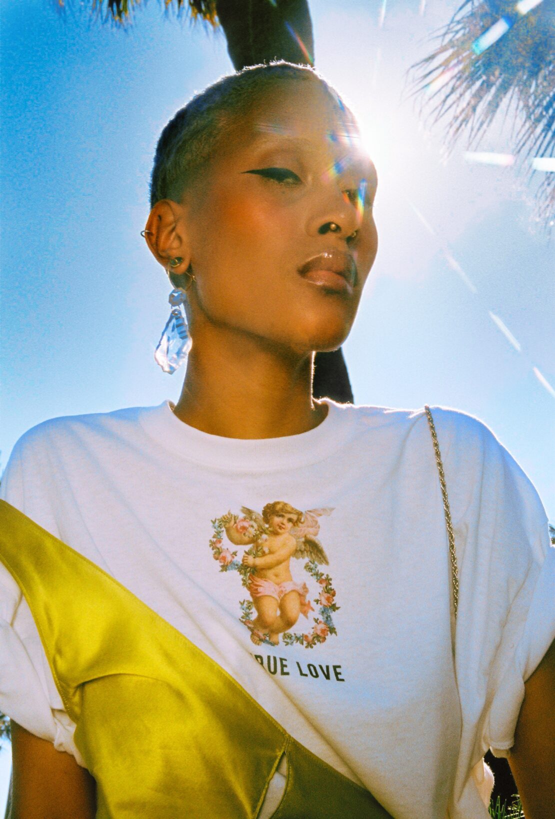 Close-up of a stylish adult female with short hair and a graphic tee standing outdoors under a palm tree in bright sunlight.