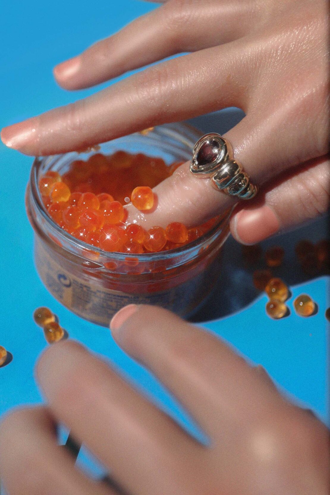 Close-up of a person's hands with a large ring on their finger, interacting with a jar of orange caviar on a blue background.