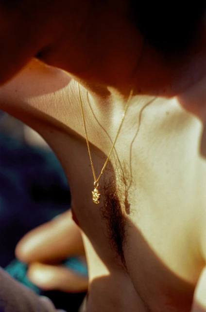 Close up of a person's neck and chest with a gold chain necklace and pendant under warm lighting, showing skin texture and detail.