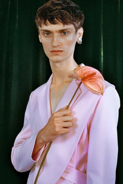 Portrait of a young adult male with short brown hair, wearing a pink suit, holding a pink Anthurium flower against a green backdrop.