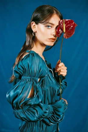 Portrait of a woman wearing a blue pleated dress holding a red anthurium flower in front of her eye against a blue background