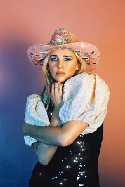 Woman in sequin dress and cowboy hat poses against a peach and blue backdrop in a studio setting with arms crossed and a serious expression.