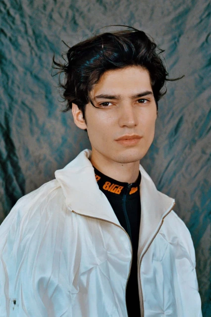 Close-up portrait of a young adult male with dark hair and fair skin wearing a white jacket against a blue patterned background.