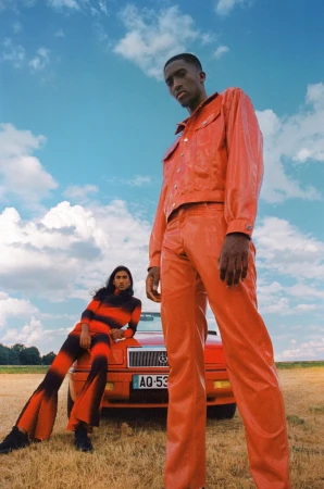 Two fashion models posing by a red vintage car in a field under a blue sky with clouds, one standing and one reclining.