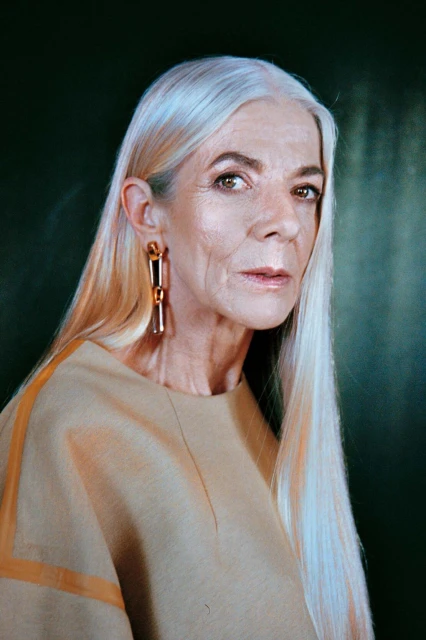 Elderly adult woman with long white hair and gold earrings posing indoors against a dark green backdrop in a portrait view.