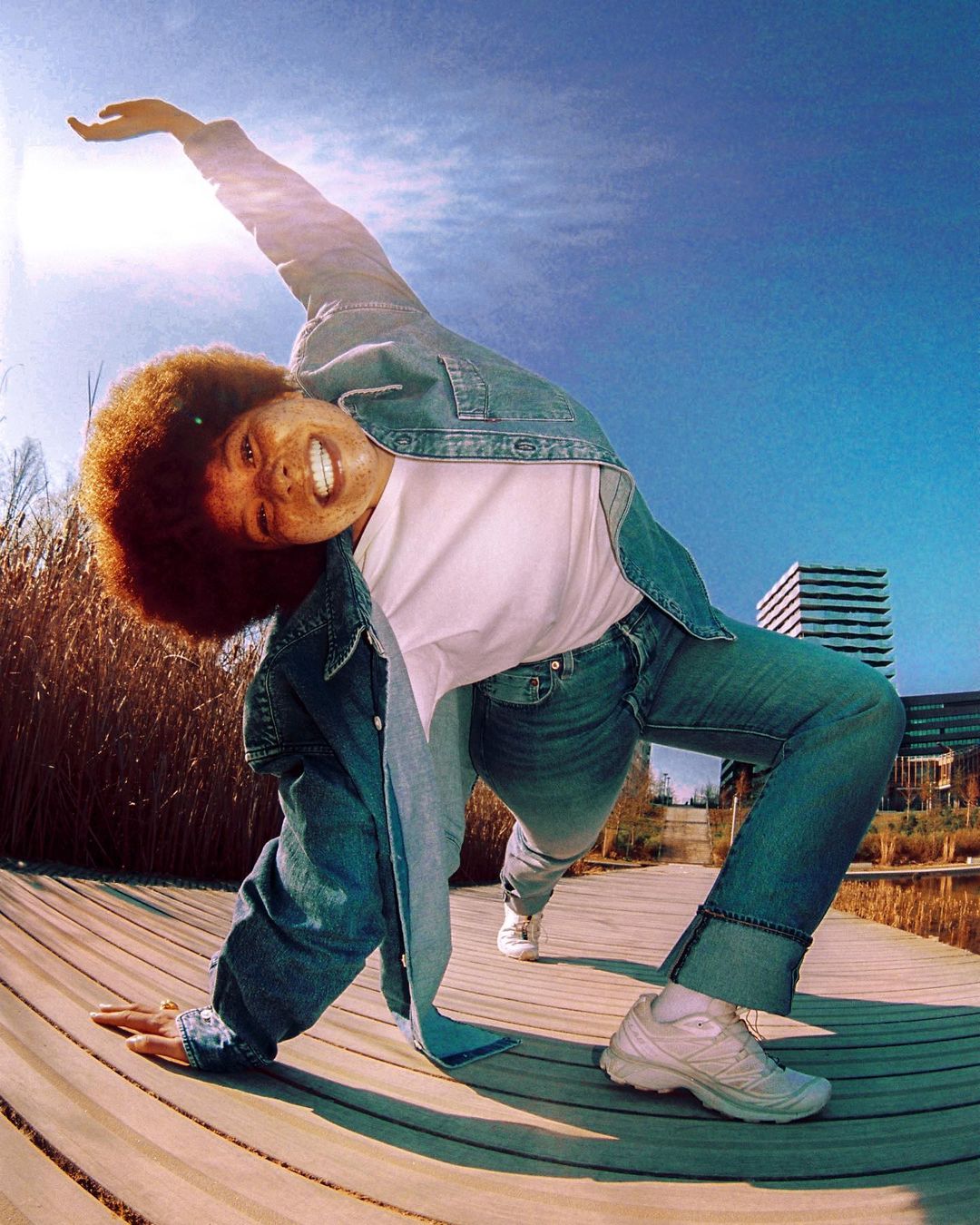 Smiling woman in denim jacket and jeans strikes yoga pose outdoors on a wooden platform under bright sunlight and a blue sky near buildings.
