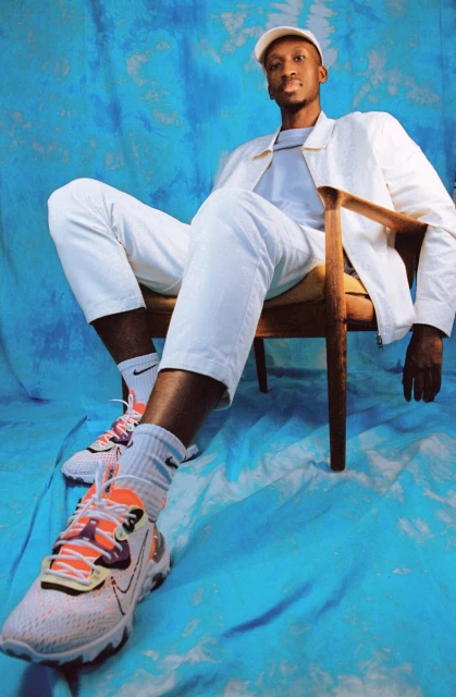 Full-length portrait of a young adult Black man wearing a white cap, jacket, pants, and colorful sneakers, seated against an abstract blue backdrop.