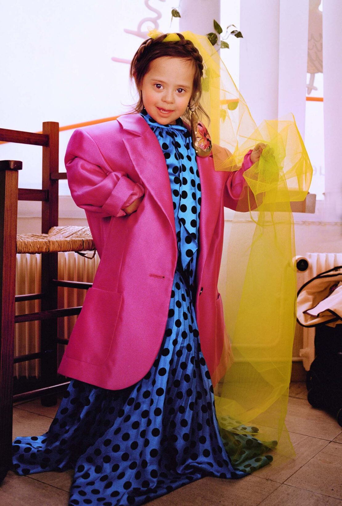 A young girl with Down syndrome smiles while wearing an oversized pink blazer and a polka dot dress with a yellow veil indoors.