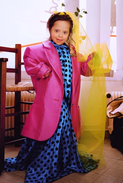 A young girl with Down syndrome smiles while wearing an oversized pink blazer and a polka dot dress with a yellow veil indoors.