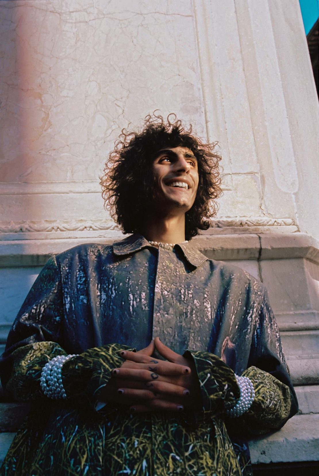 Portrait of a smiling man with curly hair wearing a decorated coat and pearl jewelry, posing against a white stone pillar outdoors.