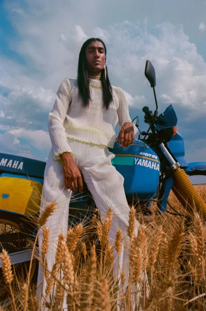 A person with long dark hair in a white pleated outfit poses with a blue motorcycle in a wheat field under a cloudy sky.