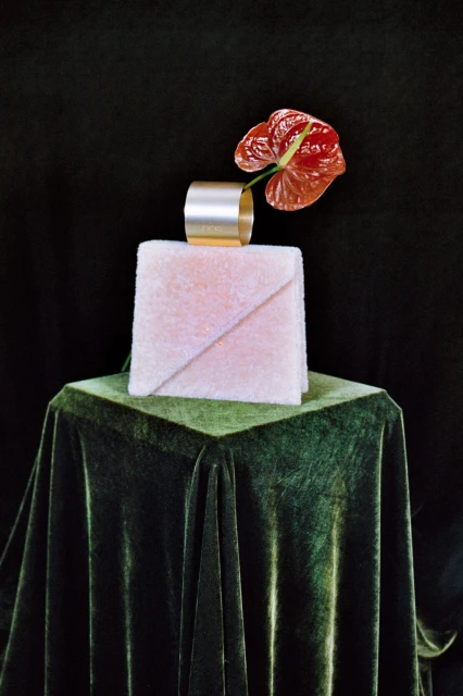 Still life shot on dark background featuring a pink textured cube topped with a gold ring and a red anthurium flower. Green velvet pedestal.