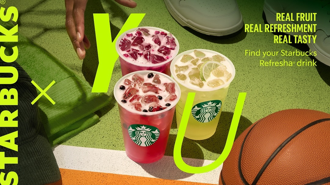 Three Starbucks Refreshers with fruit and ice in cups, next to a basketball, a hand, and a white sneaker, against a green court background.
