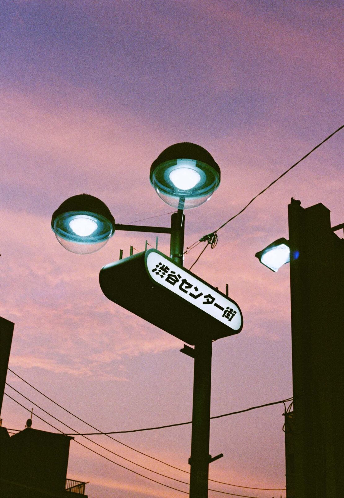 Illuminated street sign with Japanese text against a purple sky in an urban setting with buildings and power lines.