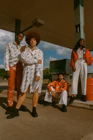 Group of four diverse young adults in retro-inspired fashion posing at a vintage gas station, captured in a medium outdoor shot.