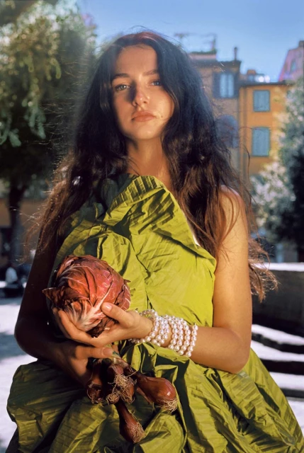 An adult woman poses in a leafy green garment, outdoors, cradling a red cabbage and red onion, wearing a pearl bracelet, with an urban background.
