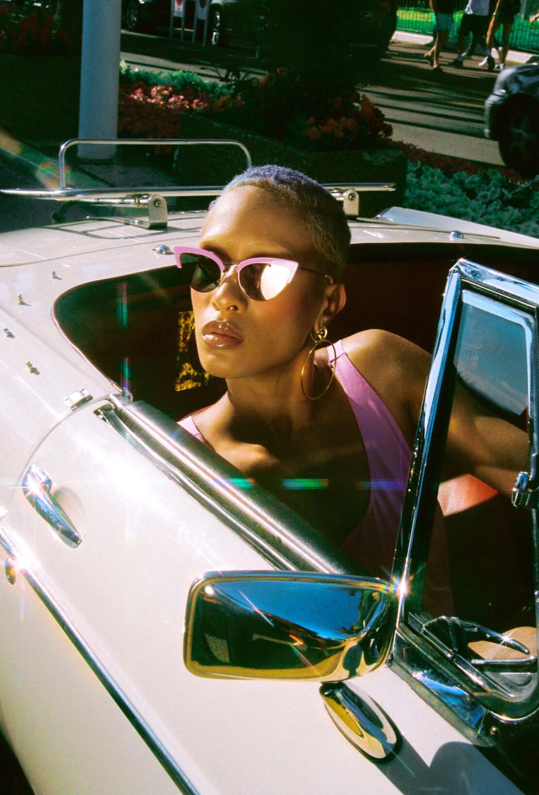 Elegant adult in a convertible, wearing pink top and sunglasses, with a gold earring, enjoying a sunny day outdoors.