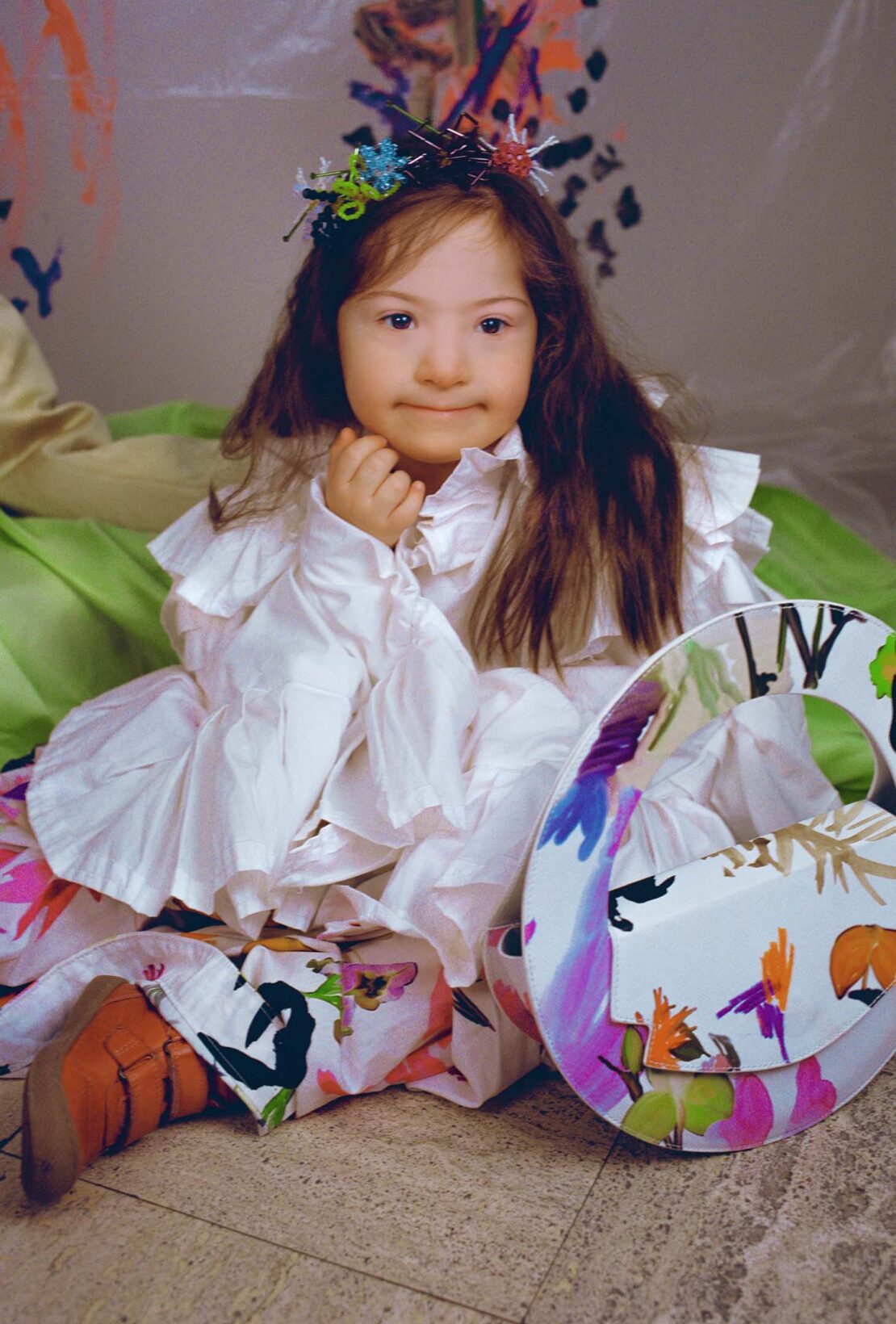 Portrait of a young girl with Down syndrome wearing a white dress and floral headpiece, sitting on the floor with painted props.