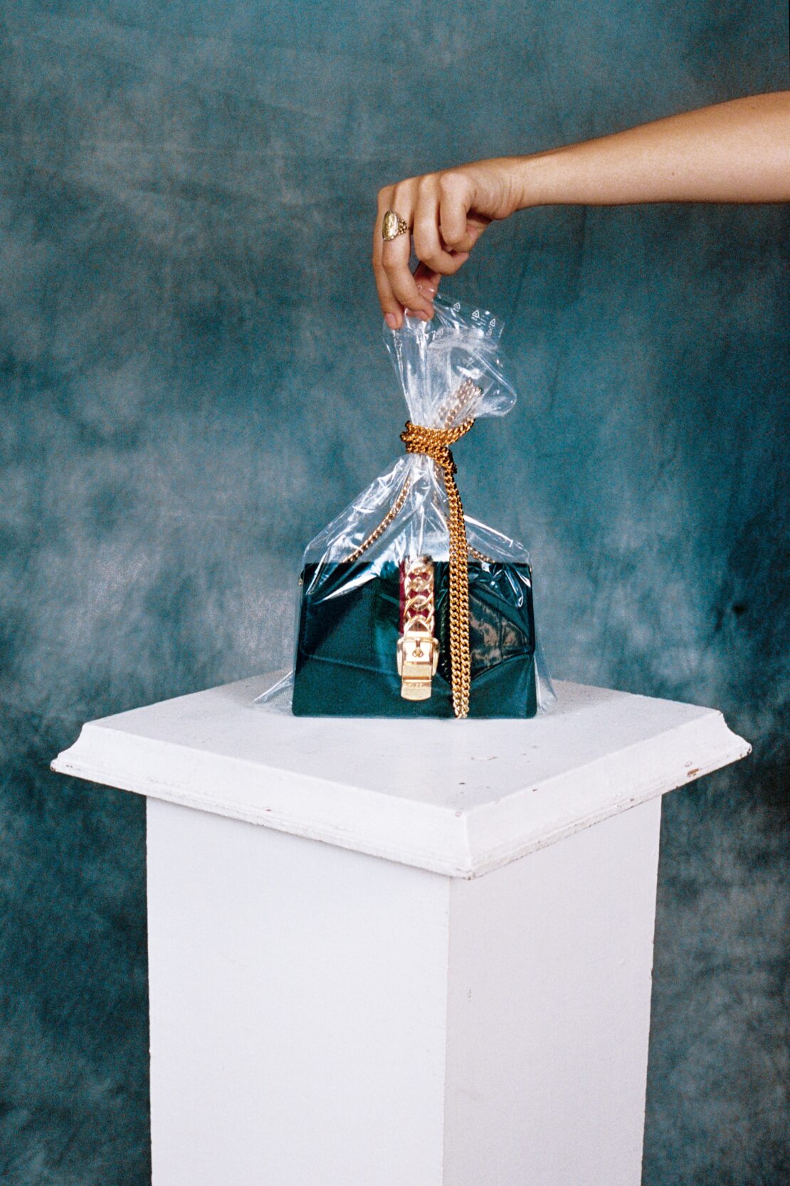 Hand holding clear plastic bag containing emerald velvet handbag with gold chain on white pedestal against mottled teal backdrop.