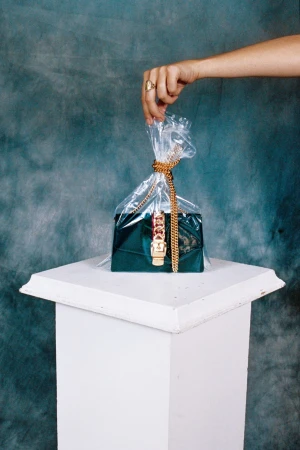 Hand holding clear plastic bag containing emerald velvet handbag with gold chain on white pedestal against mottled teal backdrop.