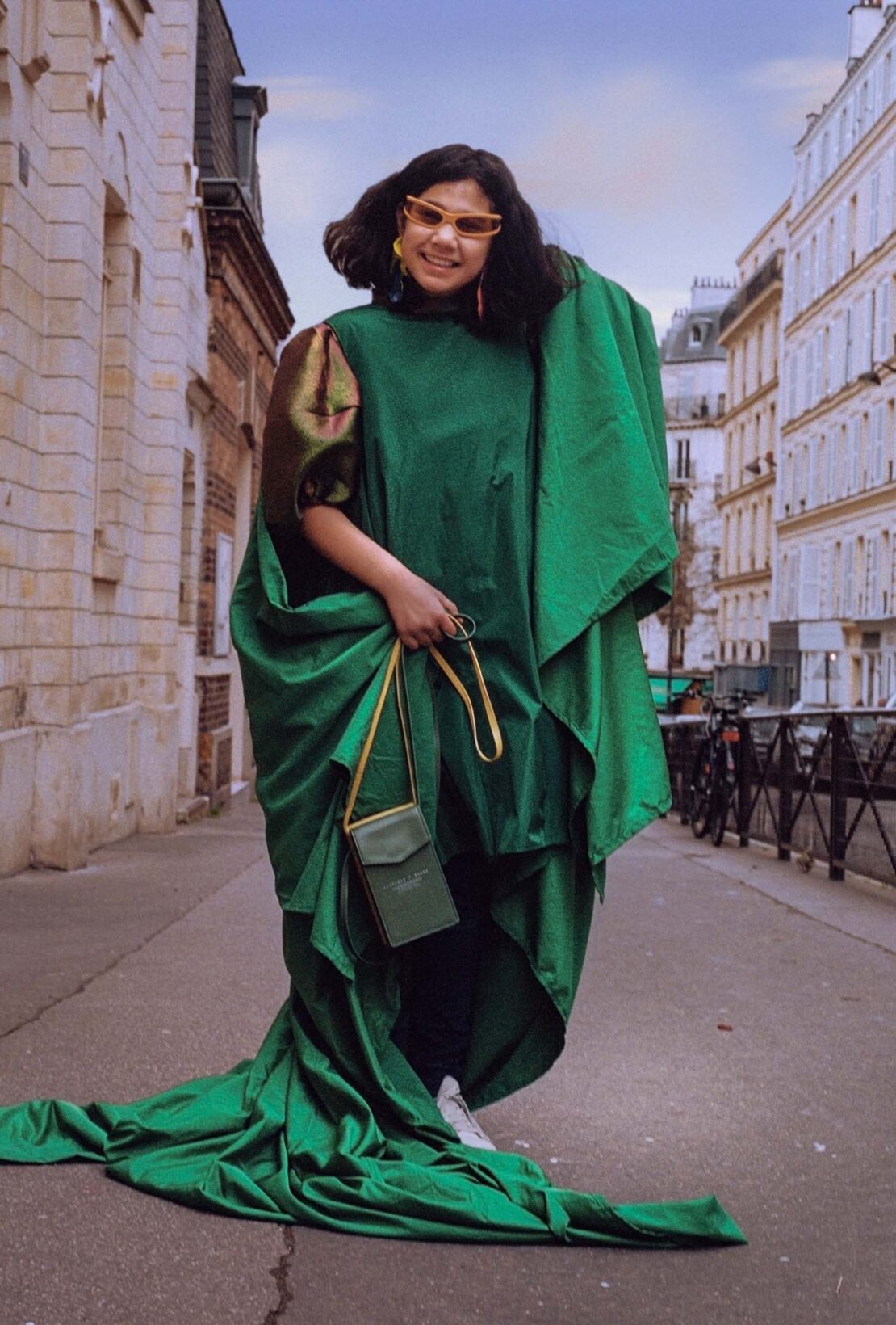 Woman in an oversized green gown wearing tinted sunglasses and holding a small crossbody bag on a European street