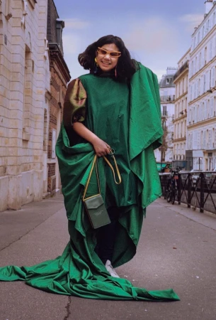 Woman in an oversized green gown wearing tinted sunglasses and holding a small crossbody bag on a European street
