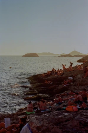 People relaxing and sunbathing on rocky coastal shorelines beside calm ocean waters during sunset light.