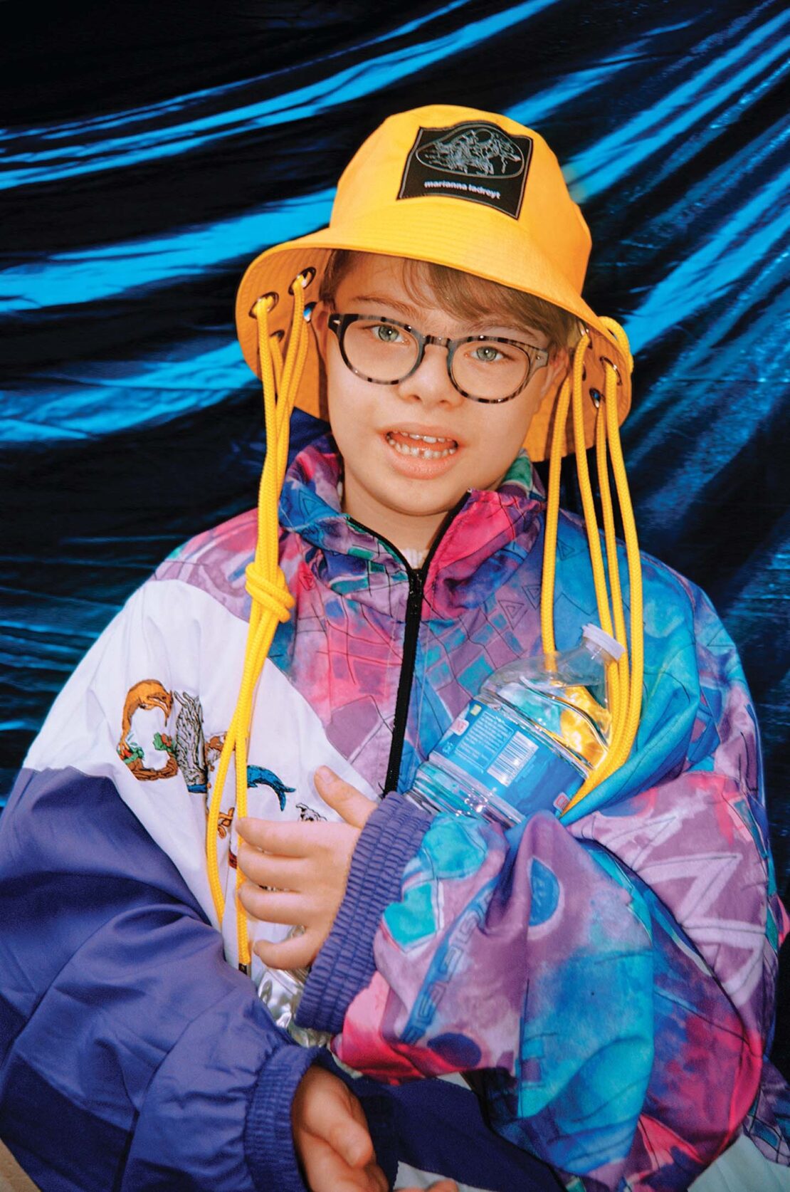 A young child with glasses wears a bucket hat and colorful jacket, holding a water bottle against a textured blue backdrop.