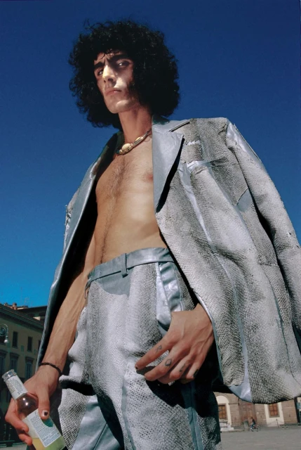 Adult male poses outdoors in a textured silver suit, holding a bottle with a blue sky and urban buildings visible in the background.