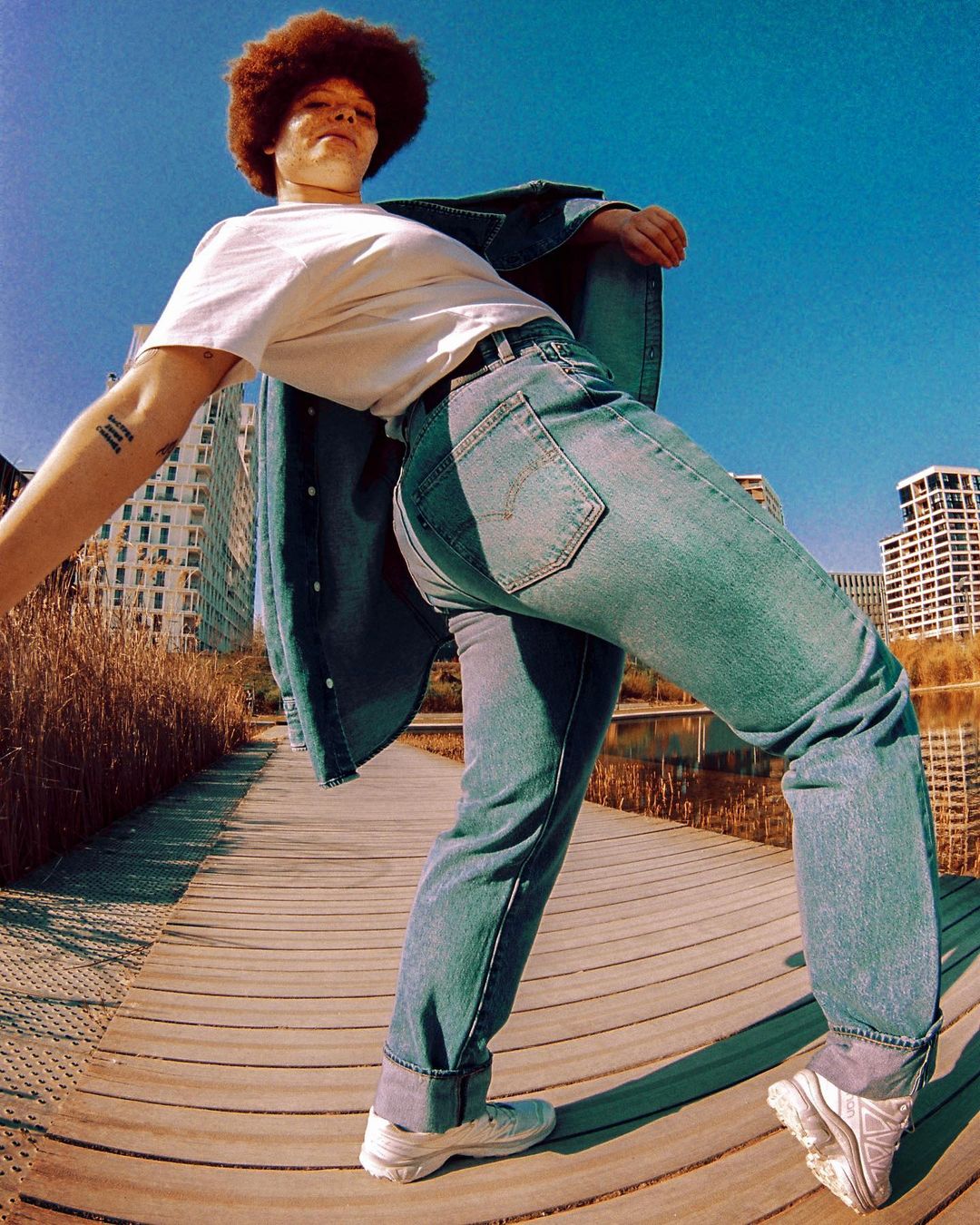 A young adult with an afro stands on a wooden boardwalk wearing jeans and a denim jacket, with buildings and the sky in the background.