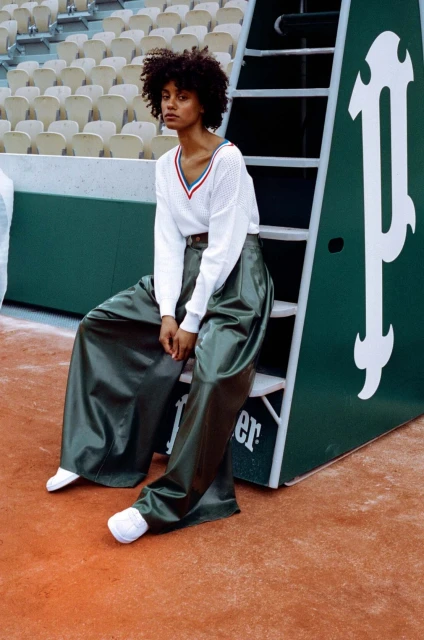 An adult female is sitting on a tennis umpire chair wearing a white sweater and green satin pants on a clay tennis court.
