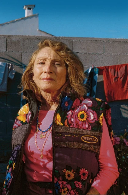 Elderly woman in floral vest poses outdoors against a concrete wall with clotheslines and a clear blue sky in the background.