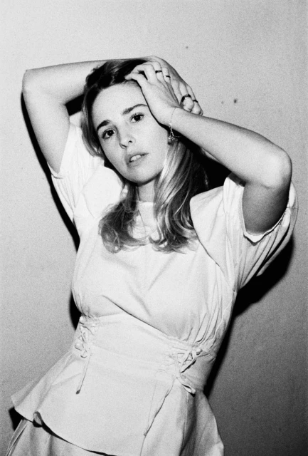 Black and white portrait of a young woman with hands on her head, wearing a white top, against a light background.