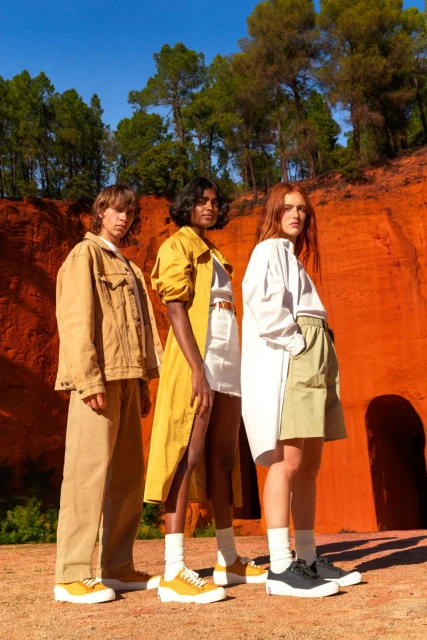 Three fashion models pose outdoors in front of red cliffs, wearing yellow, beige, and white clothing with matching sneakers and socks.