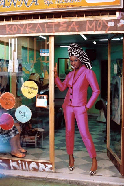 A black woman in a pink suit and zebra-print headwrap stands in the doorway of a hair salon, casting a confident gaze at the camera.