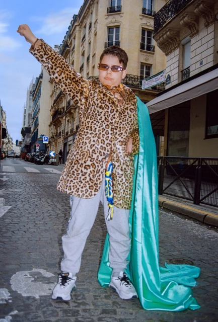 Person in leopard-print coat with blue cape in city street, standing with raised fist in an editorial fashion photograph.