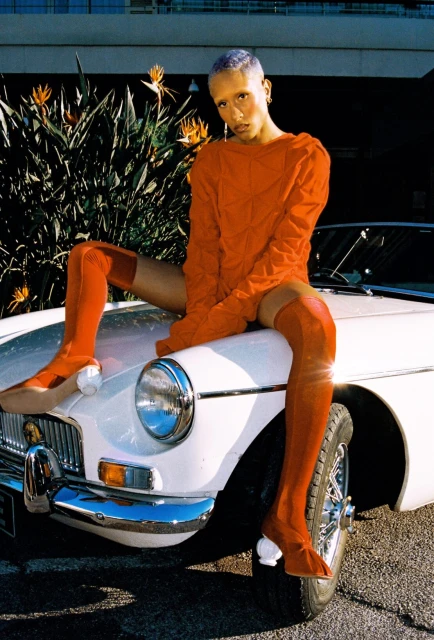 Stylish woman with orange boots poses on white vintage car in outdoor setting with tropical foliage and urban background under daylight.