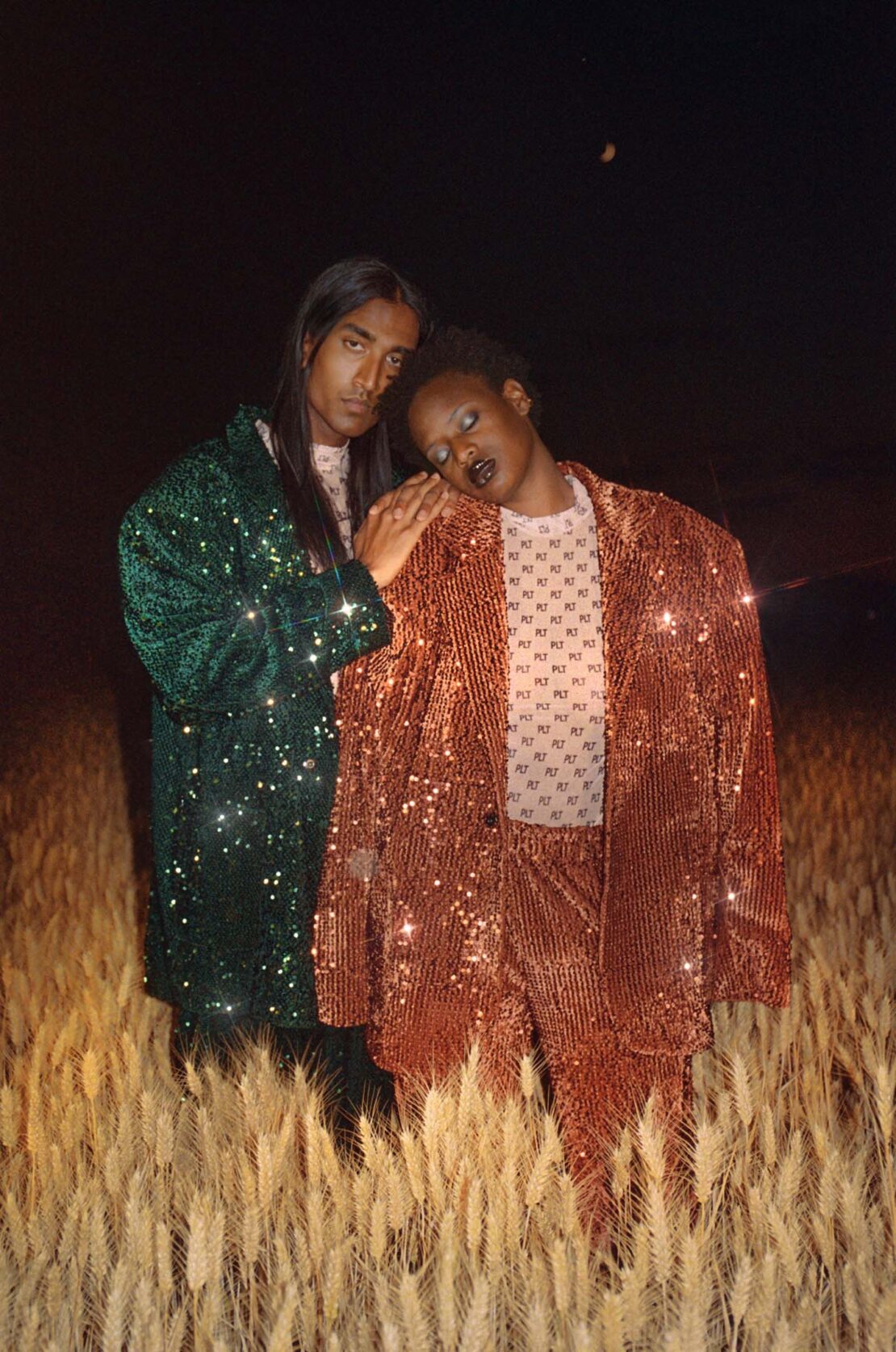 Two people stand in a wheat field at night. One person is wearing a green sequined blazer, the other a red sequined suit. Dark sky.