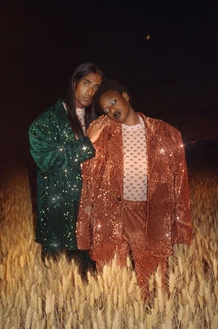 Two people stand in a wheat field at night. One person is wearing a green sequined blazer, the other a red sequined suit. Dark sky.